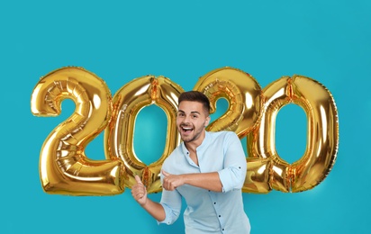 Happy young man near golden 2020 balloons on turquoise background. New Year celebration