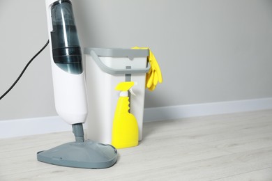 Photo of Modern steam mop, bucket with gloves and spray of cleaning product on floor near grey wall, space for text