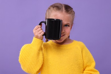Happy girl covering eye with black ceramic mug on violet background
