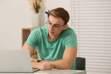 Photo of Young man with laptop writing in notebook at table indoors