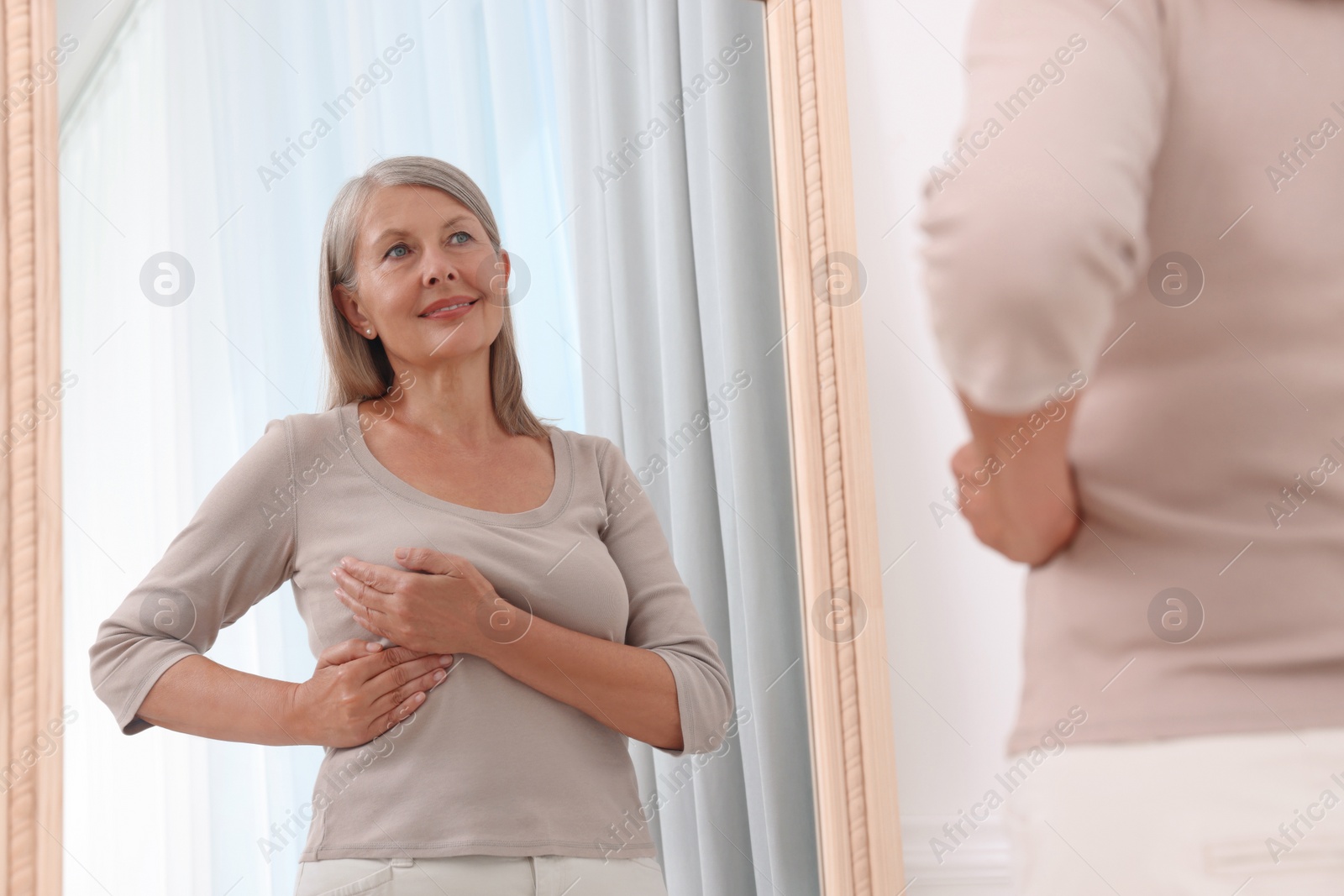 Photo of Beautiful senior woman doing breast self-examination near mirror indoors