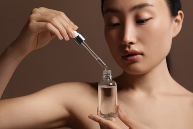 Beautiful young woman with bottle of cosmetic serum on brown background, closeup