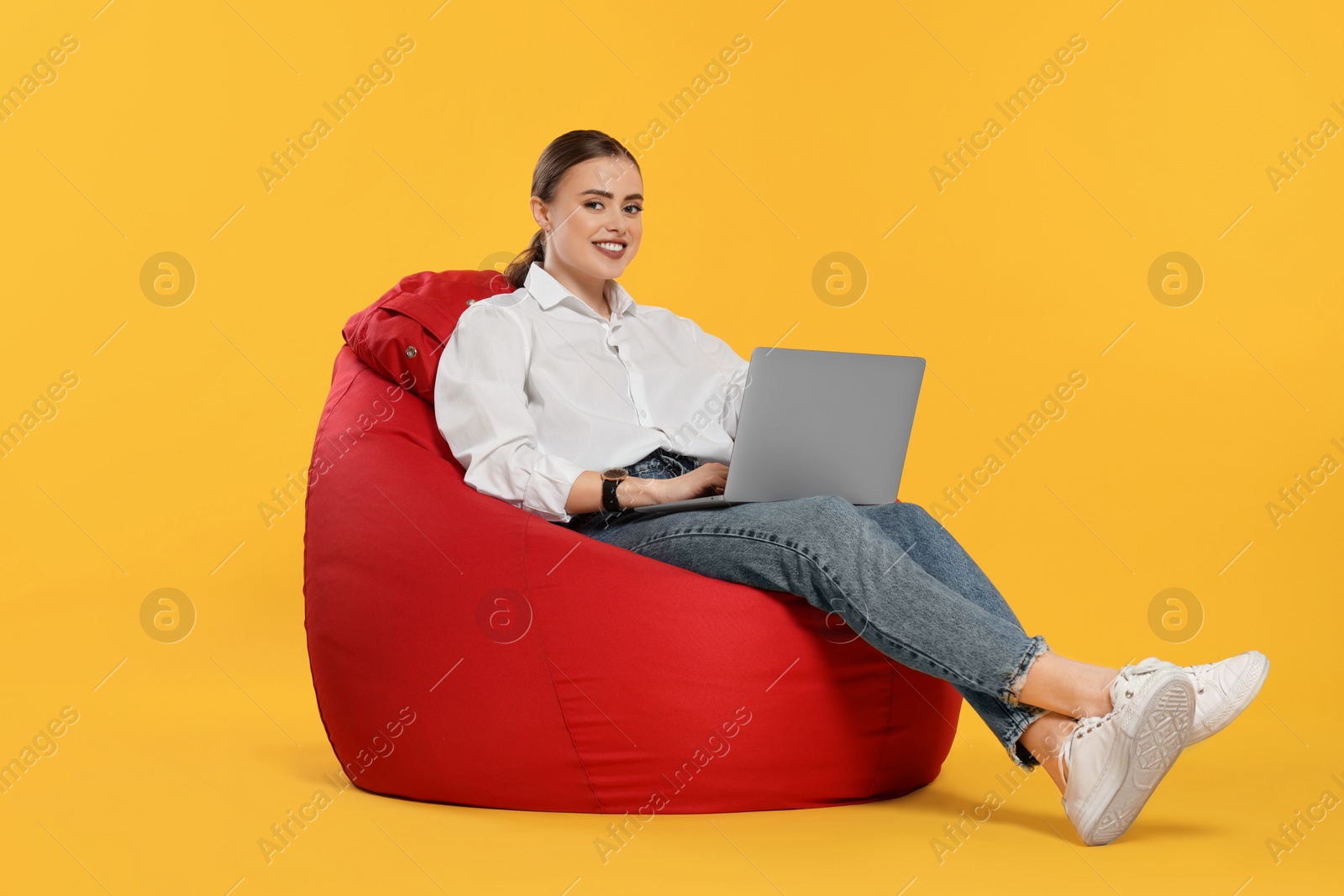 Photo of Happy woman with laptop sitting on beanbag chair against orange background