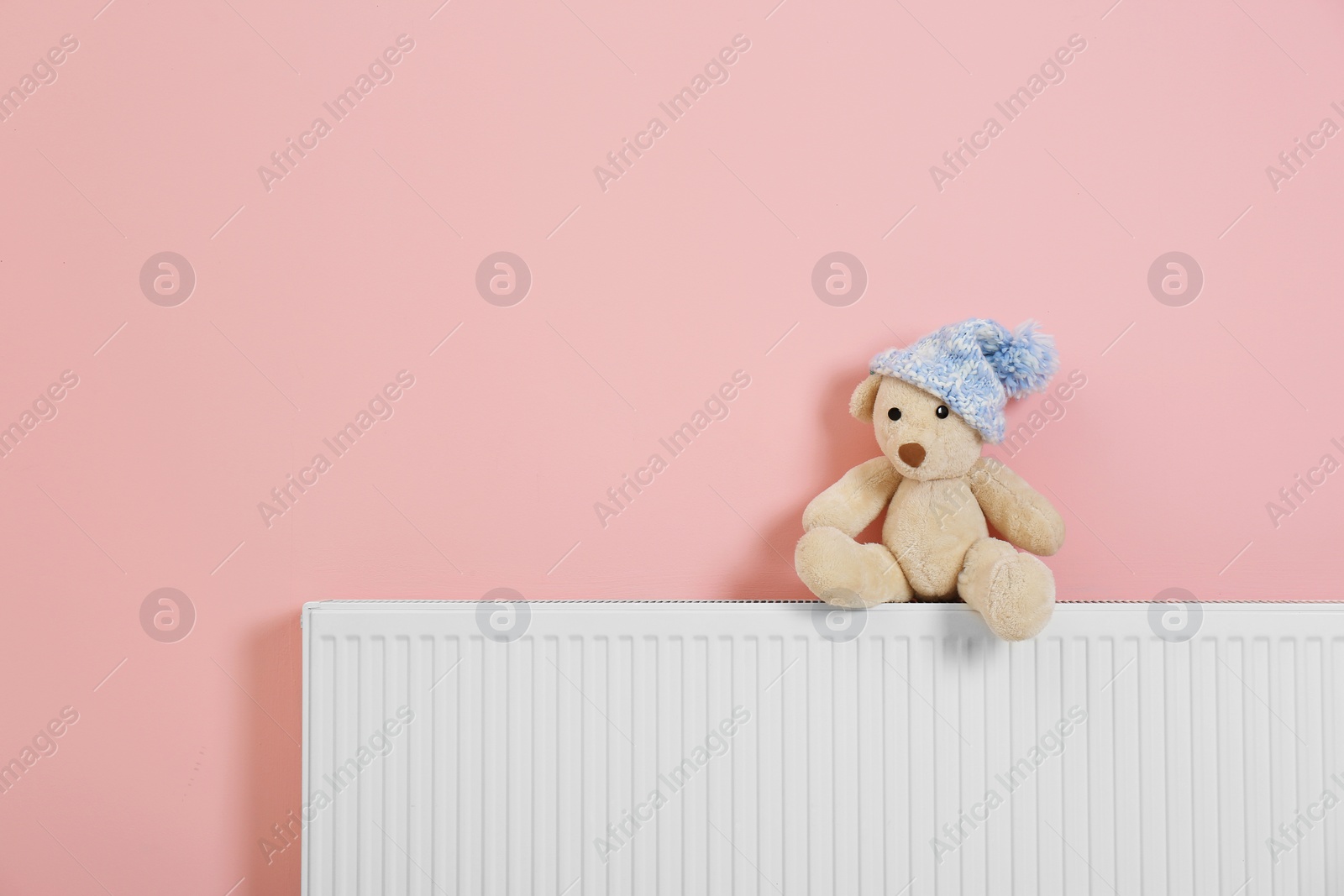 Photo of Teddy bear with knitted hat on heating radiator near color wall. Space for text