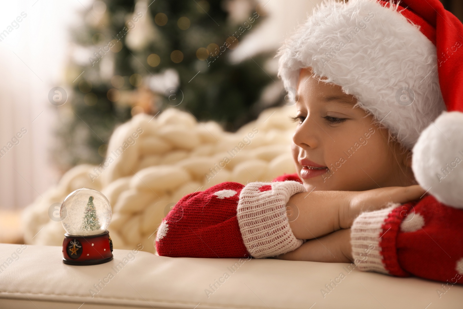 Photo of Little girl in Santa hat playing with snow globe at home