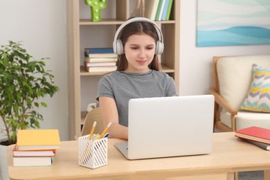 Photo of Cute girl using laptop and headphones at desk in room. Home workplace