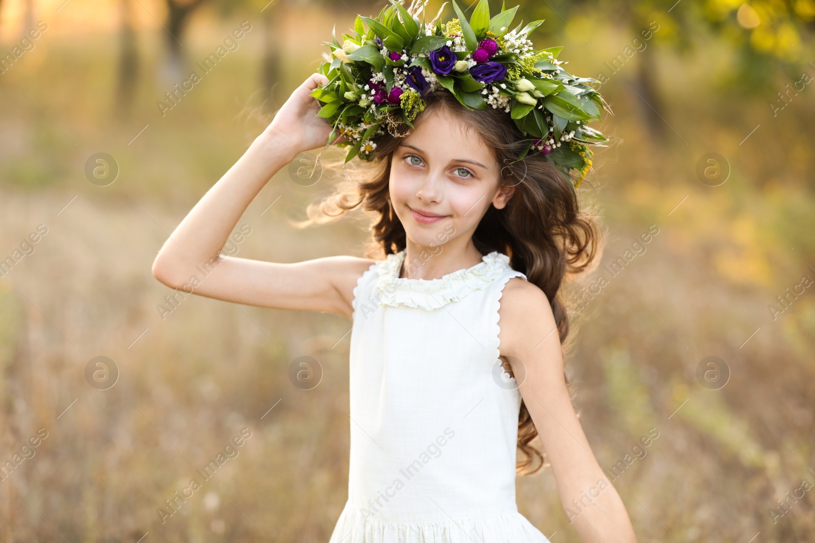 Photo of Cute little girl wearing wreath made of beautiful flowers in field on sunny day