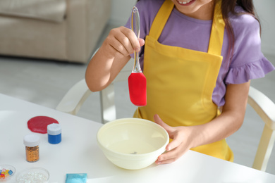 Photo of Little girl mixing ingredients with silicone spatula at table indoors, closeup. DIY slime toy
