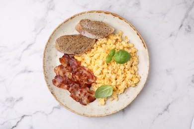 Photo of Delicious scrambled eggs with bacon and basil in plate on white marble table, top view