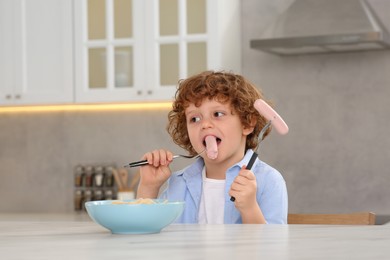 Photo of Cute little boy eating sausage and pasta at table in kitchen