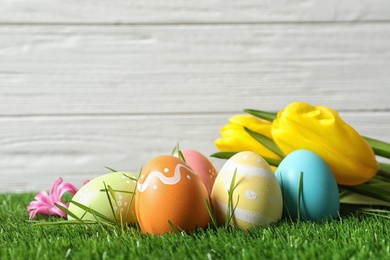 Photo of Colorful painted Easter eggs and flowers on green grass against wooden background, space for text