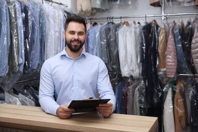 Photo of Dry-cleaning service. Happy worker holding clipboard at counter indoors