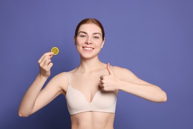 Photo of Woman in bra holding condom and showing thumb up on purple background. Safe sex