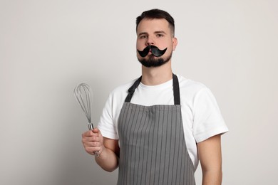 Photo of Funny confectioner in apron holding whisk on light grey background