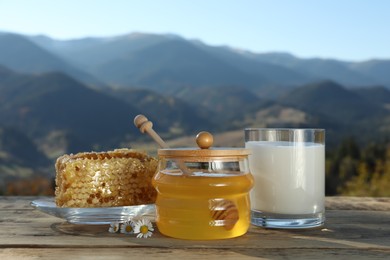 Photo of Fresh aromatic honey, combs and glass of milk on wooden table against mountain landscape