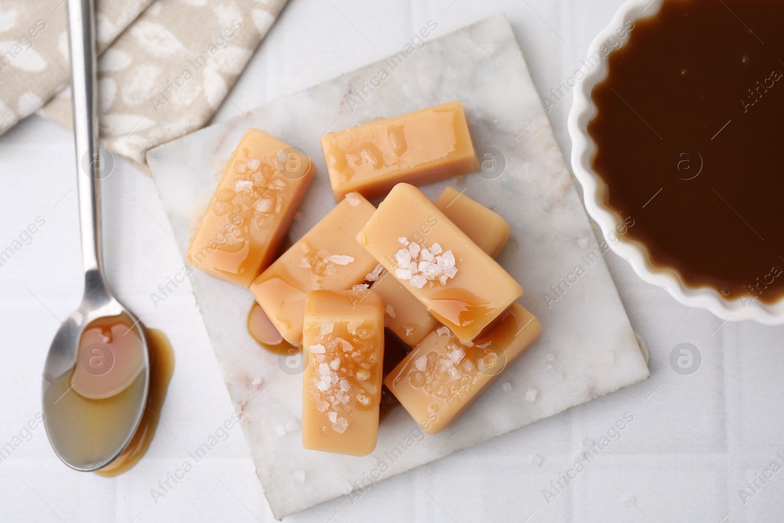 Photo of Delicious candies with sea salt and caramel sauce on white tiled table, flat lay