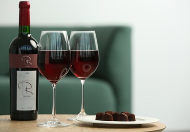 Photo of Bottle and glasses of red wine with chocolate candies on wooden table in living room