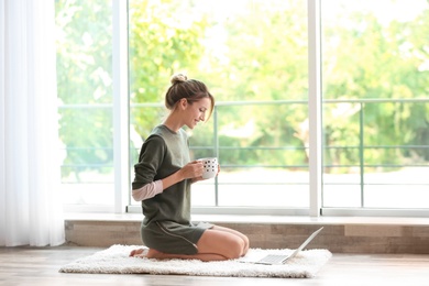 Photo of Young woman with cup of aromatic coffee and laptop sitting on floor near window at home