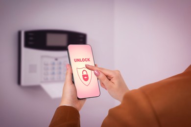 Image of Woman operating home alarm system via mobile phone against white wall with security control panel, closeup. Application interface on device screen