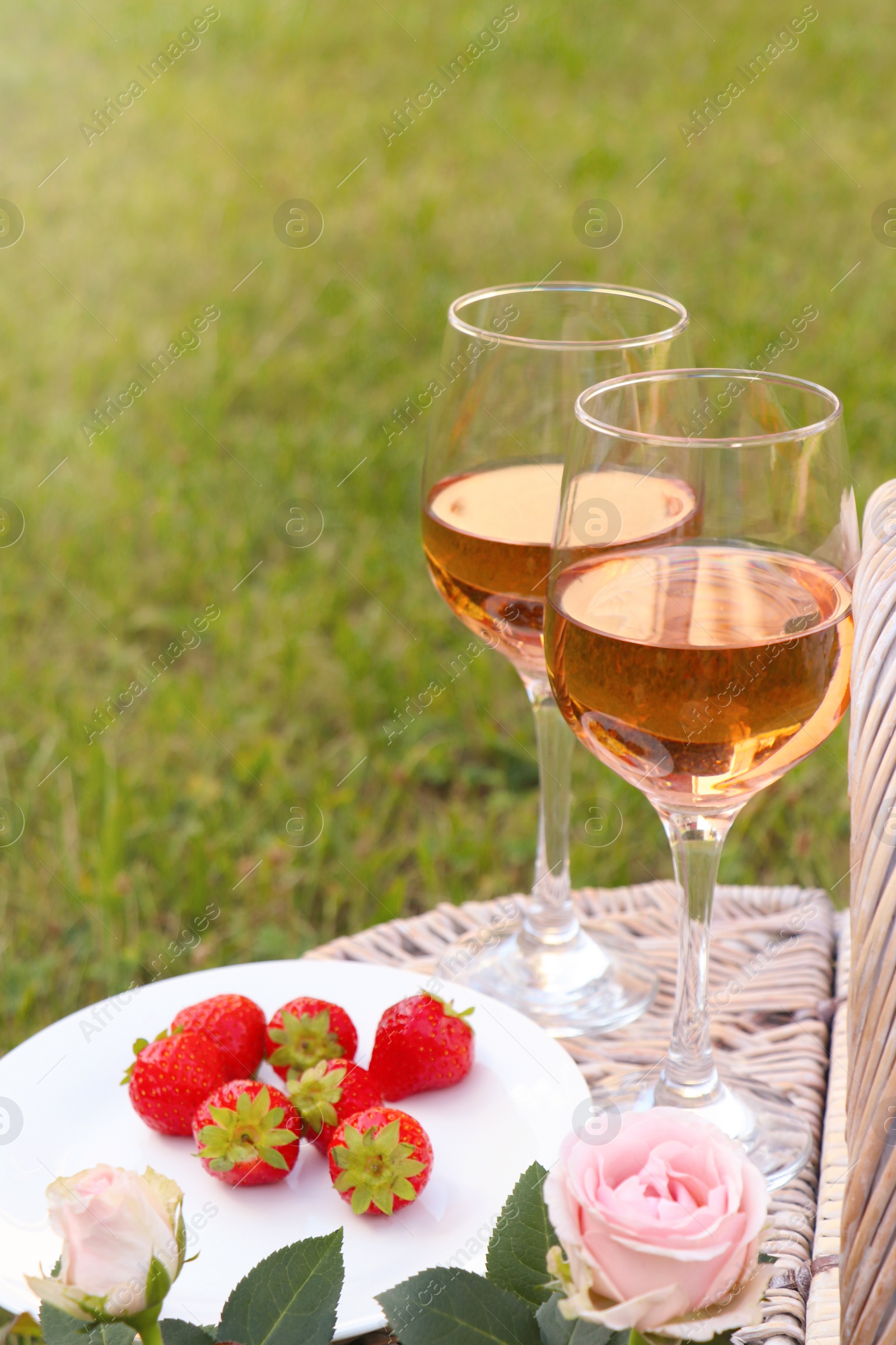 Photo of Picnic basket with glasses of delicious rose wine, strawberries and flowers outdoors