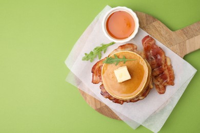 Photo of Delicious pancakes with bacon, butter, arugula and honey on green table, top view. Space for text