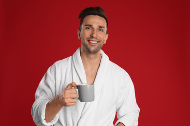 Photo of Happy young man in bathrobe with cup of coffee on red background