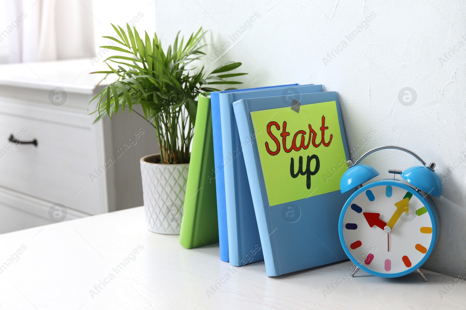 Photo of Book with words Start Up, plant and alarm clock on white wooden table indoors