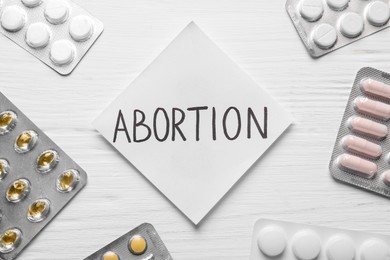 Photo of Paper note with word Abortion and pills on white wooden background, flat lay