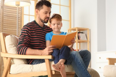 Father reading book with his son on armchair in living room at home