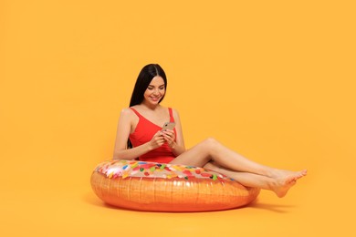 Photo of Happy young woman with beautiful suntan using smartphone on inflatable ring against orange background