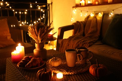 Rattan furniture, cups, fairy lights, burning candles and other autumn decor on outdoor terrace at night