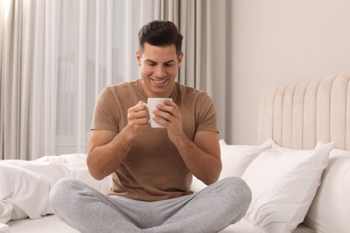 Happy man with cup of hot drink on bed at home