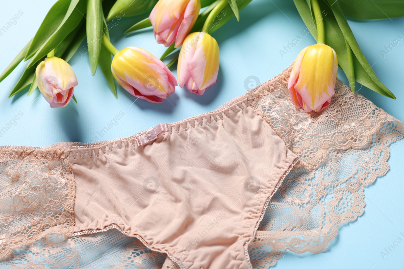 Photo of Elegant beige women's underwear and beautiful tulips on light blue background, closeup