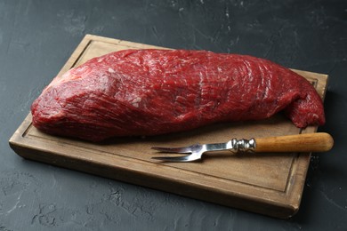 Photo of Wooden board with piece of raw meat and fork on black table