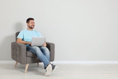 Happy man with laptop sitting in armchair indoors, space for text
