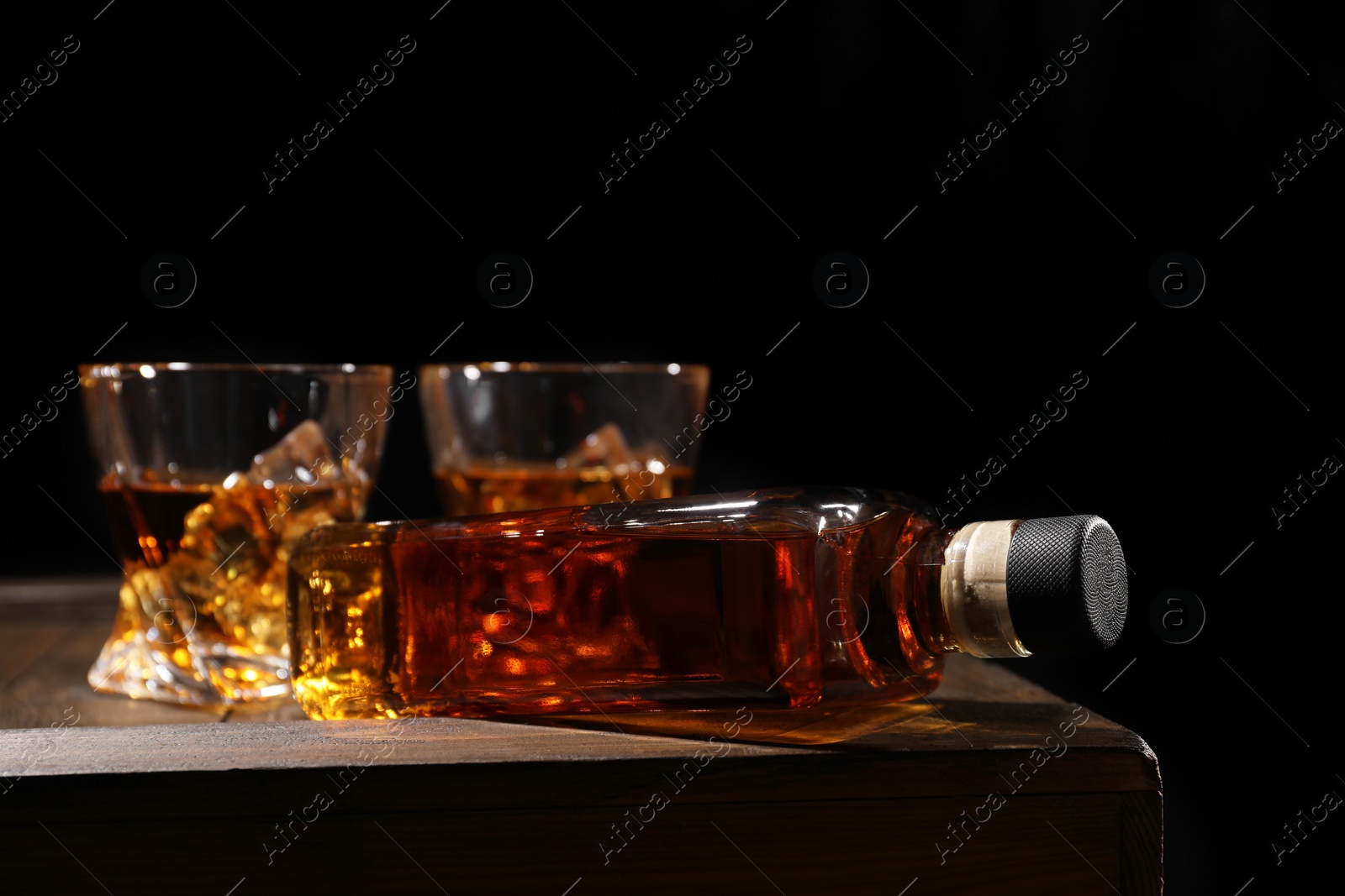 Photo of Whiskey with ice cubes in glasses and bottle on wooden crate against black background, closeup