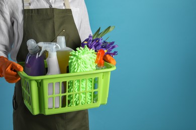 Spring cleaning. Woman holding basket with detergents, flowers and tools on light blue background, closeup. Space for text