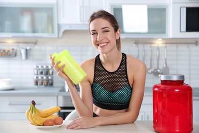 Photo of Young woman holding bottle of protein shake at table with ingredients in kitchen