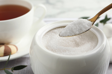 Photo of Spoon with granulated sugar over bowl on table, closeup