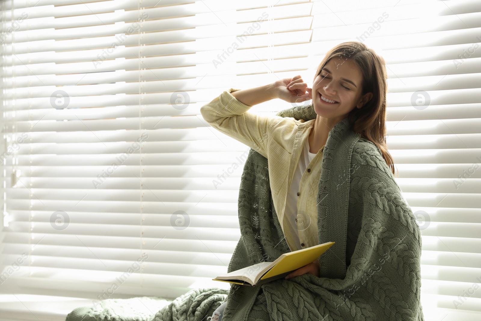 Photo of Woman covered with warm green plaid reading book near window indoors