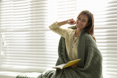 Photo of Woman covered with warm green plaid reading book near window indoors