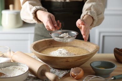 Making dough. Woman sifting flour into bowl at wooden table in kitchen, closeup