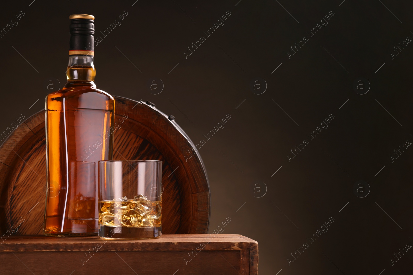 Photo of Whiskey with ice cubes in glass and bottle on wooden table near barrel against dark background, space for text