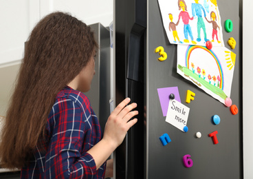 Woman opening refrigerator door with child's drawings, notes and magnets in kitchen