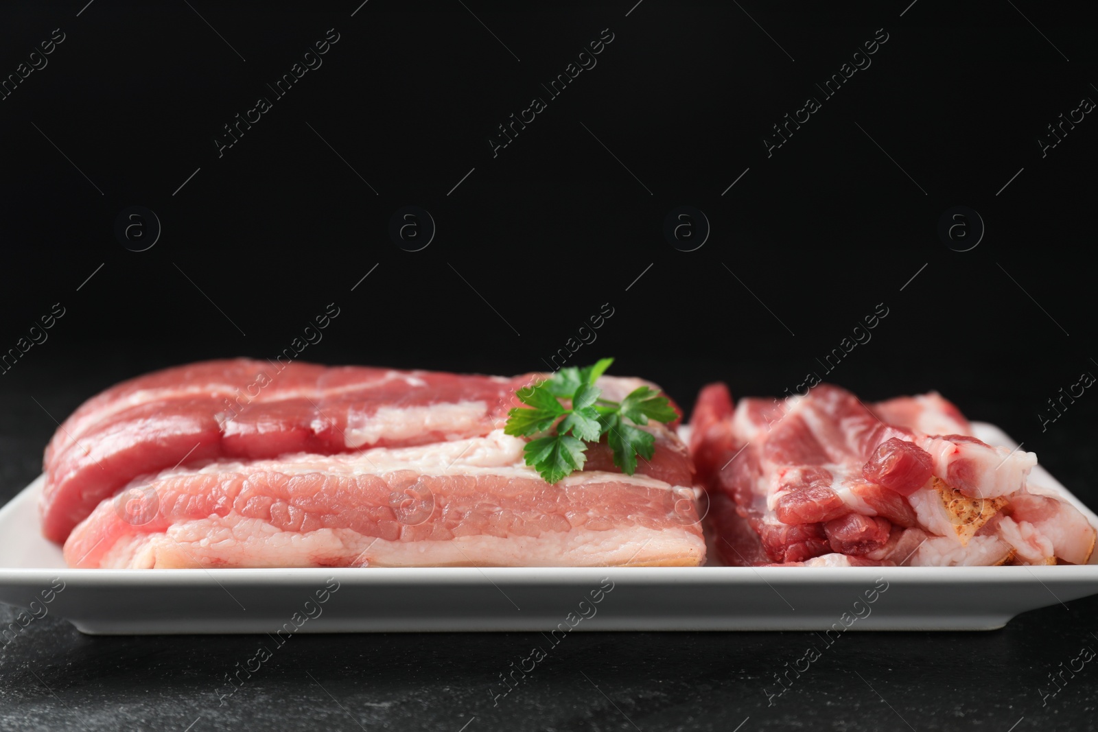 Photo of Pieces of raw pork belly and parsley on black textured table, closeup. Space for text