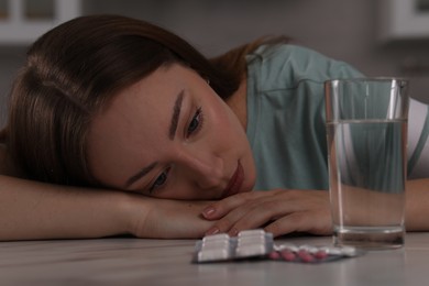 Depressed woman at table with antidepressant pills and glass of water indoors