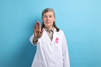 Photo of Mammologist with pink ribbon showing stop gesture on light blue background. Breast cancer awareness