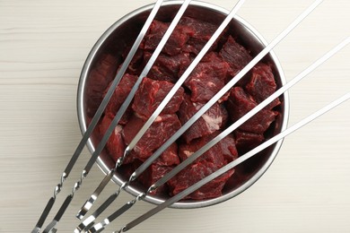 Metal skewers and pot with raw meat on white wooden table, top view