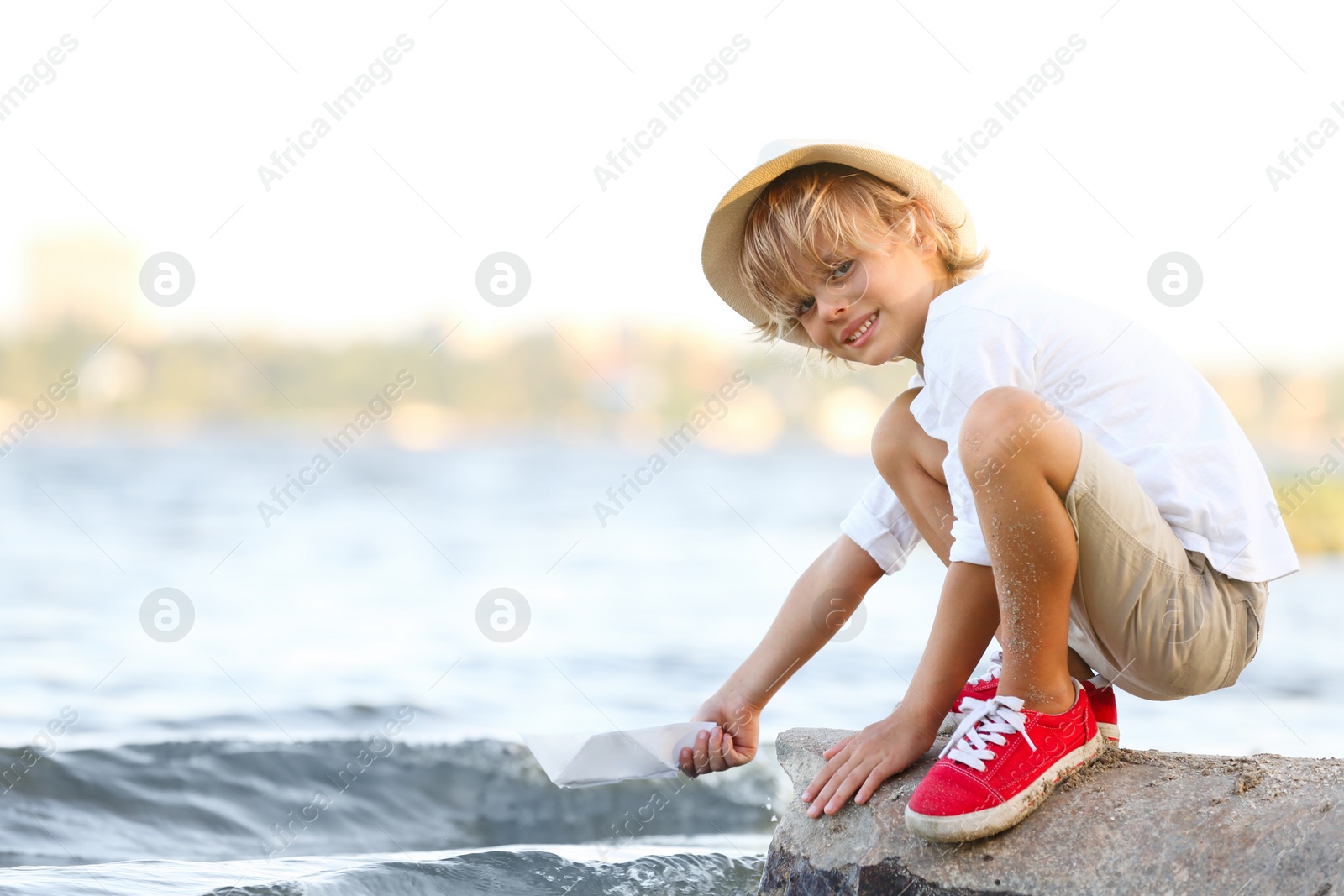 Photo of Cute little boy with paper ship at river, space for text. Child spending time in nature