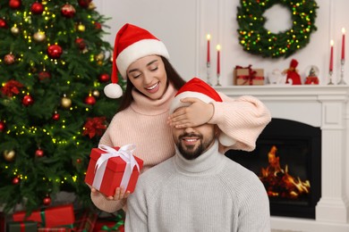 Happy woman in Santa hat surprising her boyfriend with Christmas gift at home